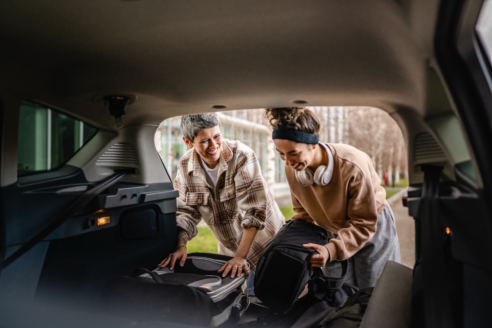 A mother and her college-age daughter laughing together while loading luggage into a car trunk.