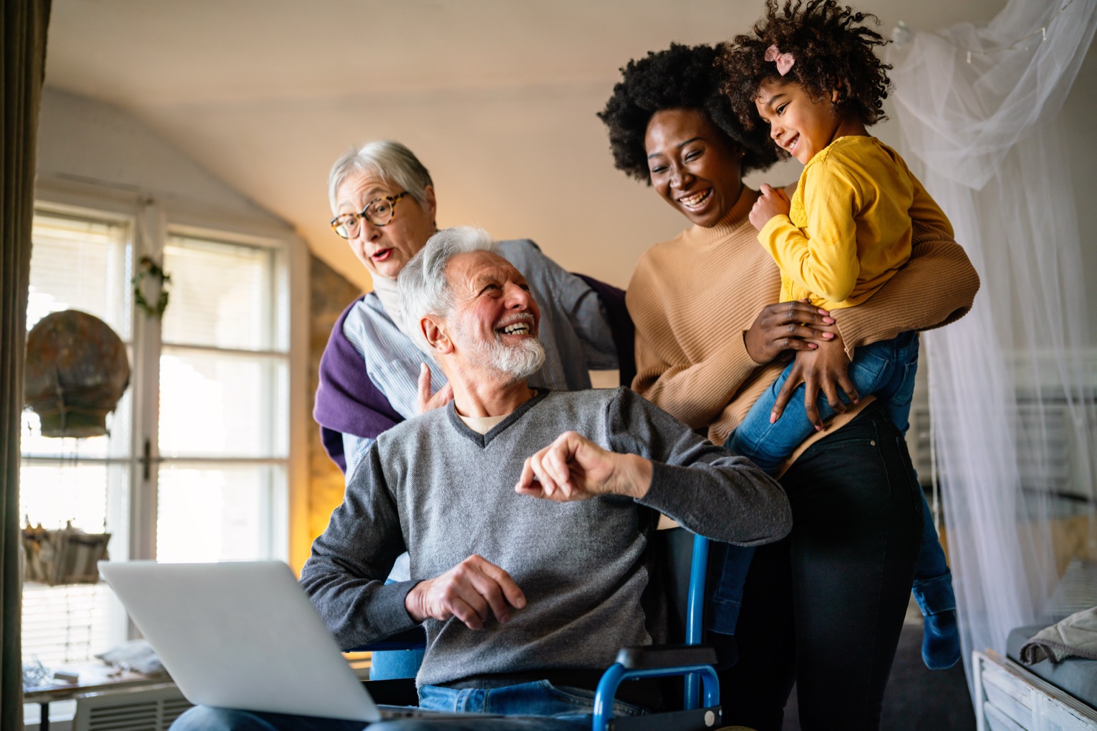 A multigenerational family gathered around a laptop for a video call, with a grandfather in a wheelchair pointing at the screen.