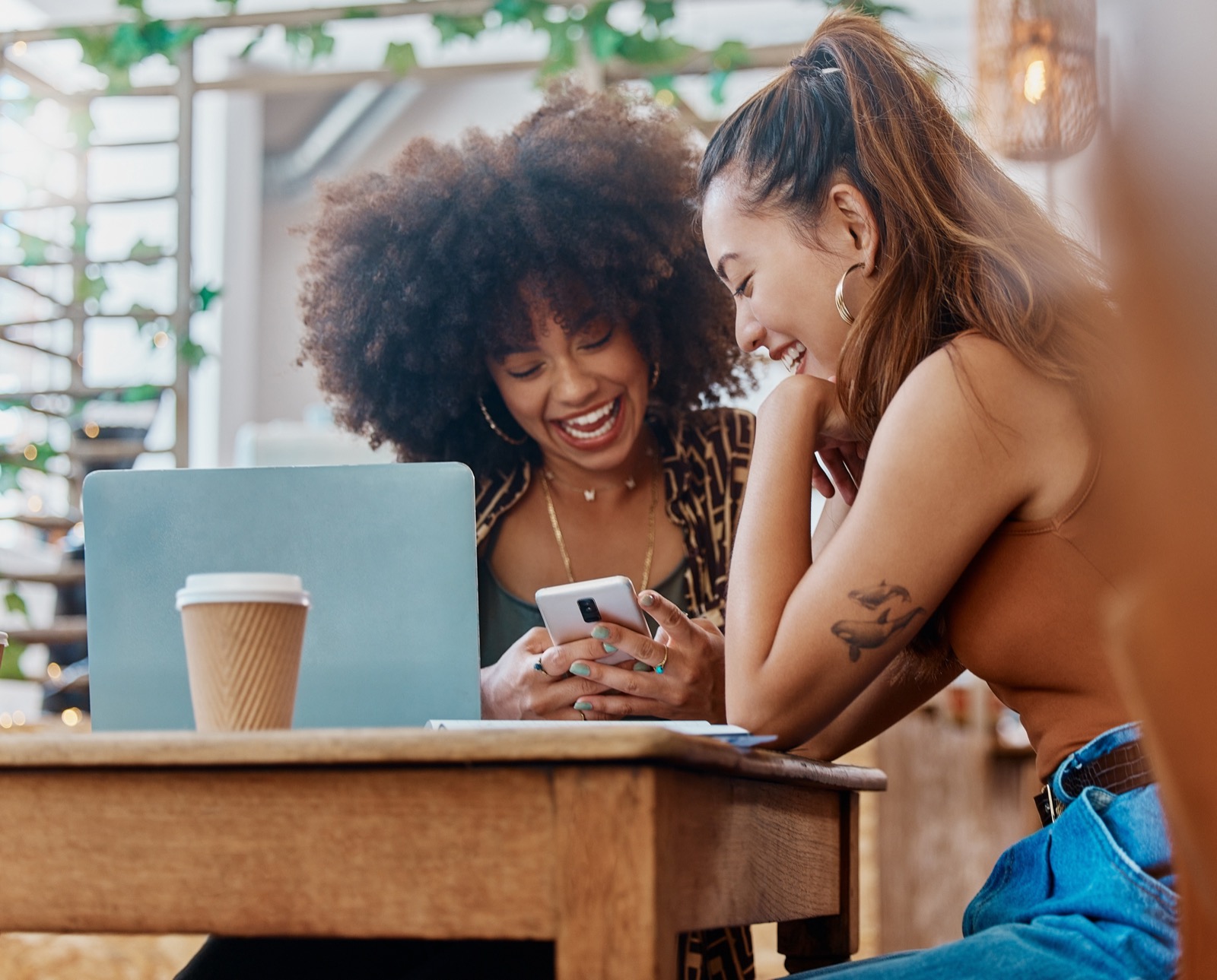 Two friends laughing together over a phone at a cafe table with a laptop and coffee.