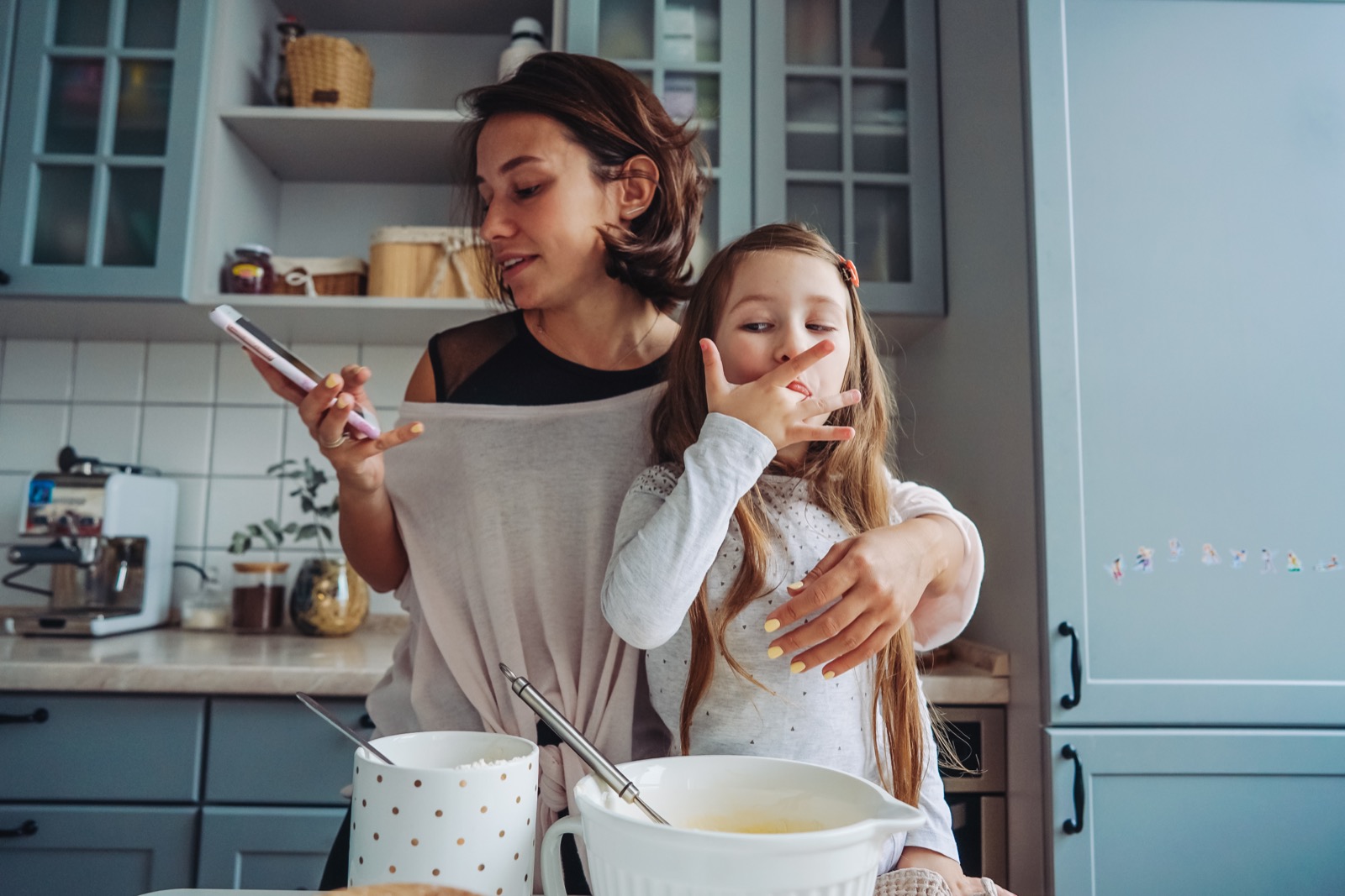 A mother checking her phone while baking in the kitchen with her young daughter.