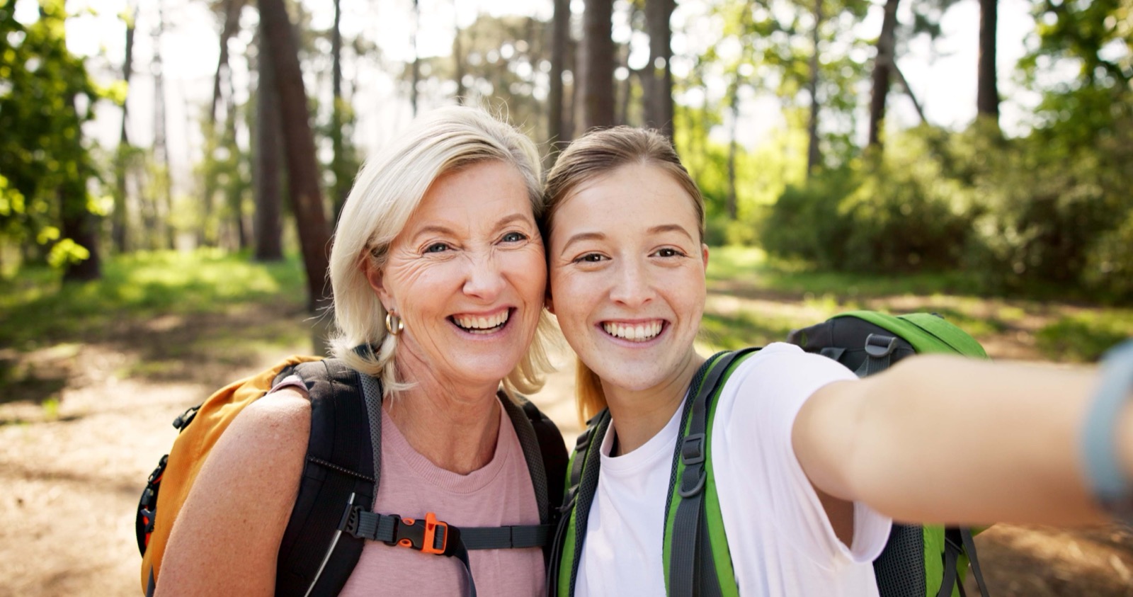 A mother and her adult daughter smiling together while hiking, both wearing backpacks.
