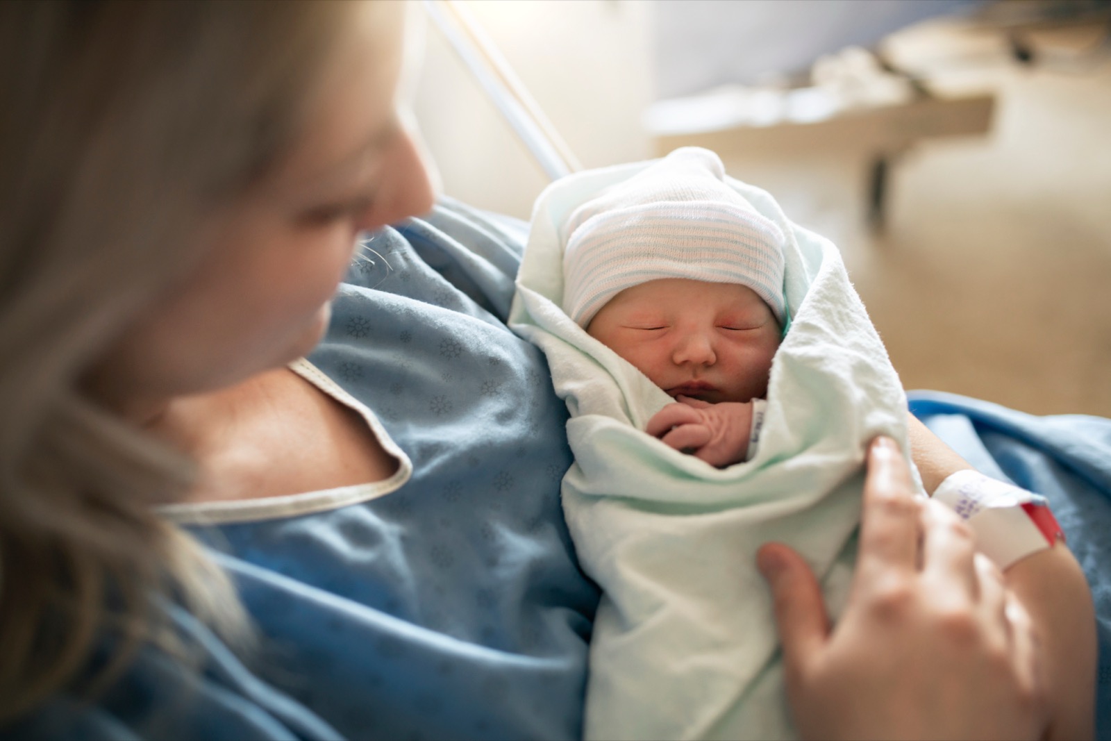 A mother in a hospital gown holding her sleeping newborn, who is wrapped in a blanket and wearing a striped hat.