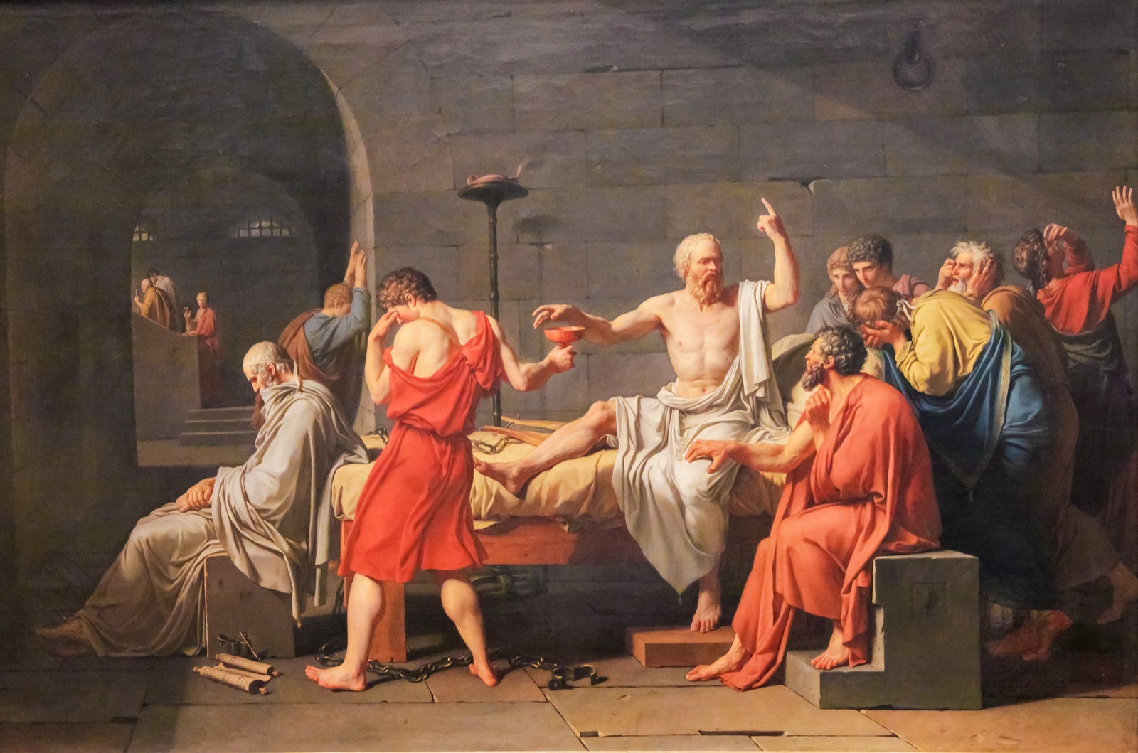 A dramatic painting of Socrates, surrounded by grieving followers, raising a hand while holding a cup of hemlock.